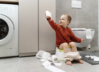 toddler in red shirt holding toilet paper and sitting on small potty learning to potty train