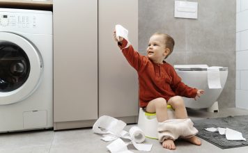 toddler in red shirt holding toilet paper and sitting on small potty learning to potty train