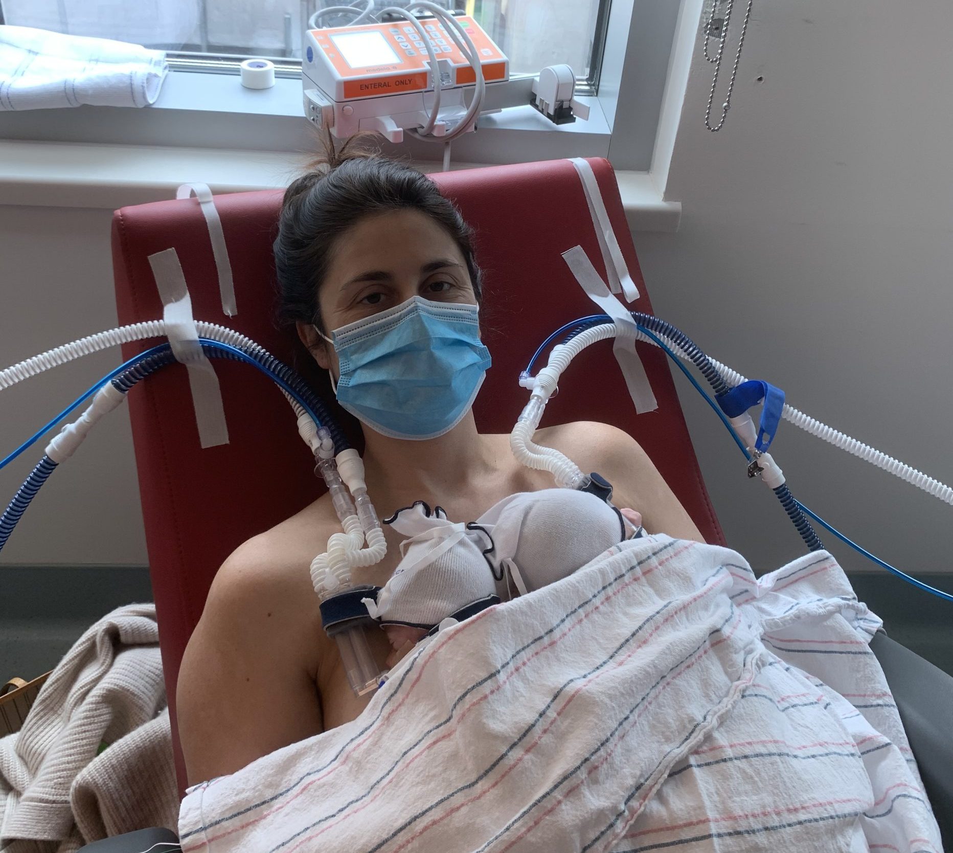 A mom wearing a face mask while holing premature babies in a chair connected to multiple medical devices.